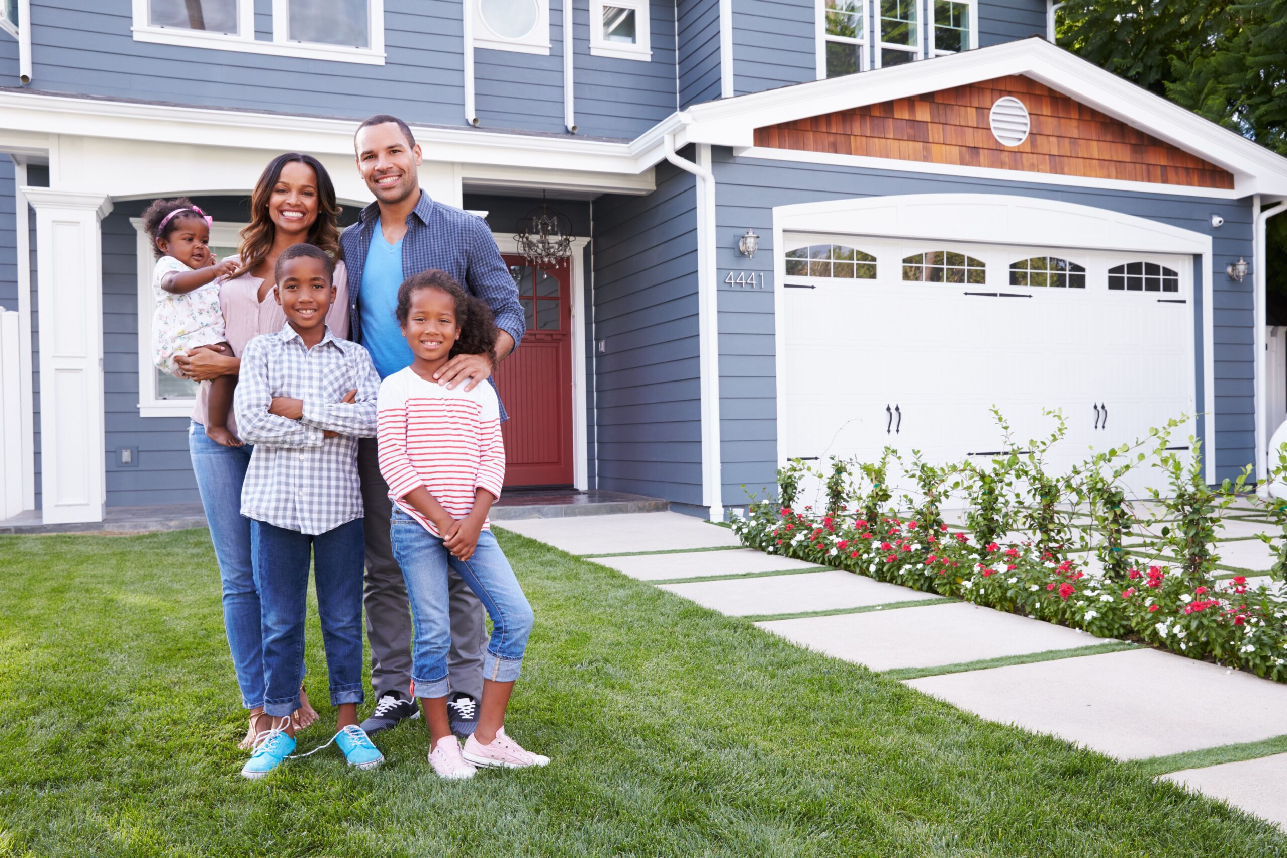 A smiling family stands in front of their modern home, with two adults and three children, showcasing joy and togetherness in a vibrant setting.