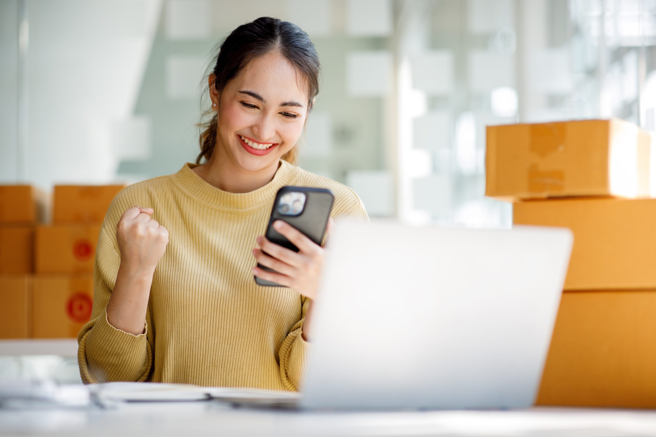 Smiling woman in a cozy sweater shows excitement while looking at her phone, with boxes and a laptop in the background, suggesting a productive workspace.