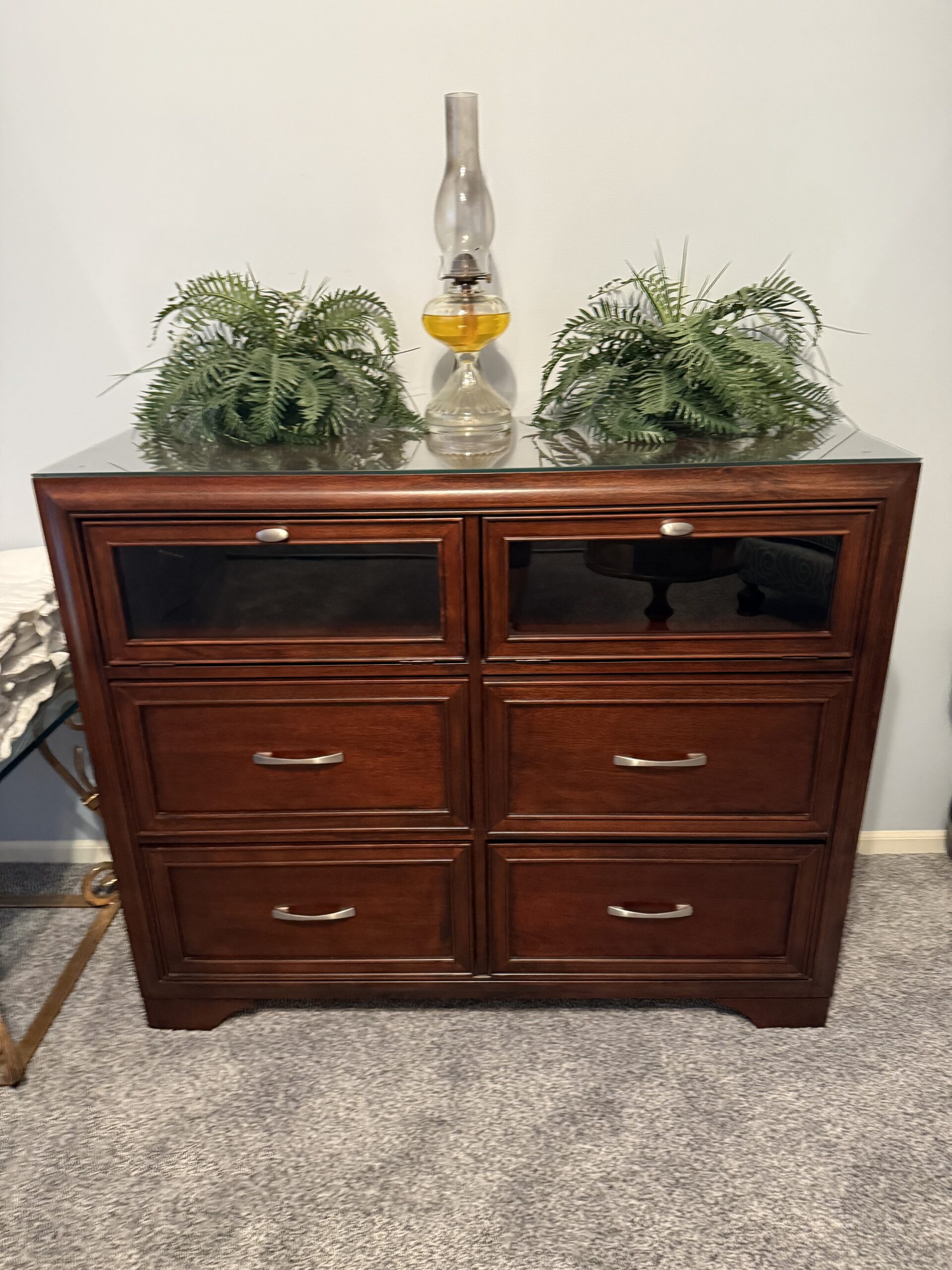 Elegant wooden dresser featuring six drawers, topped with a vintage oil lamp and two decorative ferns, set against a neutral wall.